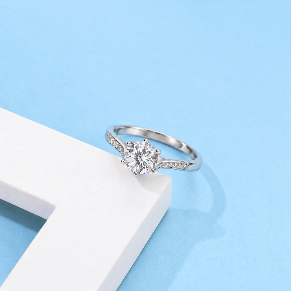 Round moissanite ring with pavé shoulders on a white prop against a pale blue background.